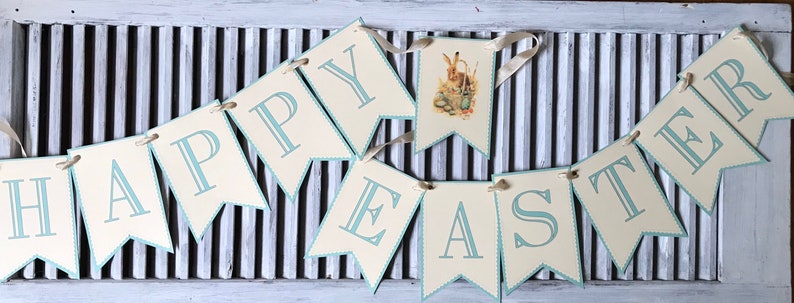 May include: A banner with the words "Happy Easter" in blue lettering on a cream background. The banner is strung with white ribbon and features a small illustration of a bunny rabbit.