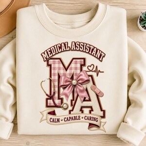 Peut inclure: Un sweat-shirt à col rond couleur crème avec un motif comprenant le texte MEDICAL ASSISTANT et les lettres MA. Le design inclut un motif à carreaux vichy rose, un nœud en satin, un stéthoscope, un pansement, un tracé cardiaque et une bannière avec le texte CALM CAPABLE CARING.