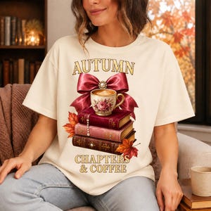 Peut inclure: Un t-shirt couleur crème avec une impression graphique représentant trois livres empilés, une tasse à thé fleurie et un grand nœud bordeaux. Le texte indique AUTUMN au-dessus du nœud et CHAPTERS & COFFEE sous les livres, avec des feuilles d'automne.