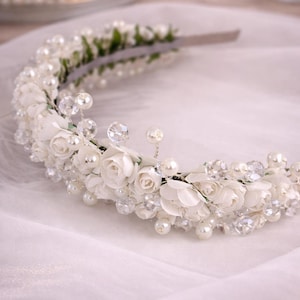 Könnte beinhalten: Ein Braut-Haarreif mit weißen Stoffrosen, Kunstperlen und klaren Kristallakzenten. Der Blumenkranz ist auf einem silberfarbenen Band angebracht. Der Haarschmuck ist für Hochzeiten und besondere Anlässe gedacht.