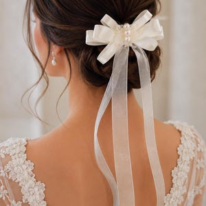 Könnte beinhalten: Ein Haaraccessoire bestehend aus einem cremefarbenen Satinband, das zu einer mehrschichtigen Schleife gebunden ist. Die Mitte der Schleife ist mit drei kleinen, runden, weißen Kunstperlen verziert. Zwei lange, transparente Bänder hängen herab.