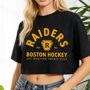 May include: Black cropped t-shirt with the text "RAIDERS BOSTON HOCKEY EST. NINETEEN TWENTY-FOUR" in yellow. The shirt features a yellow emblem with a letter "R" inside. The shirt is worn by a person.