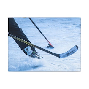 Può includere: Primo piano di un giocatore di hockey su ghiaccio in azione. Si vedono le gambe del giocatore con pantaloni neri e pattini, con una mazza da hockey. La mazza ha una lama curva. Ghiaccio e trucioli di ghiaccio sono visibili.