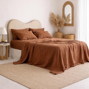 May include: A bed with rust-colored linen bedding, including a fitted sheet, flat sheet, and pillowcases. The bed is against a neutral-toned headboard. A woven rug and a woven pendant light are also visible.