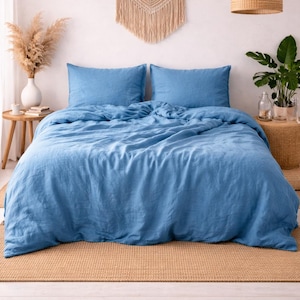 Pode incluir: Um conjunto de roupa de cama de linho azul numa cama, incluindo uma capa de edredão e duas fronhas. A cama está sobre um tapete tecido, com uma tapeçaria de macramé por cima. Uma lâmpada pendente tecida pende do teto.