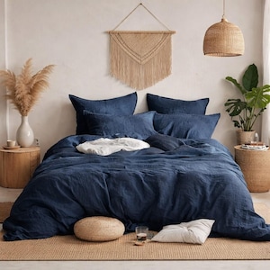 May include: A navy blue linen bedding set on a bed, including a duvet cover and pillows. The bed is on a woven rug, with a white pillow and a small woven ottoman at the foot of the bed. A macrame wall hanging and a woven pendant light are visible.