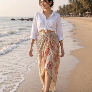 May include: A white long-sleeved button-down shirt tied at the waist and a beige sarong skirt featuring a repeating pattern of small dark blue floral motifs and larger red tree-shaped designs with a draped, asymmetrical hemline.