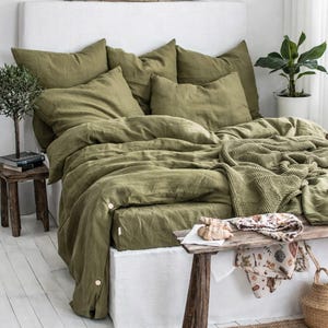 Peut inclure: Un lit avec une parure de lit vert olive, comprenant une housse de couette et des oreillers, ainsi qu'une couverture en tricot. Le lit est adossé à une tête de lit blanche. Une petite table en bois contient des livres et une plante. Un tapis tissé et un banc sont devant le lit.