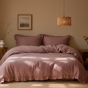 May include: A bedroom scene featuring a bed with a dusty rose-colored duvet cover and matching ruffled pillowcases. The bed is the focal point, complemented by a wooden side table, a small stool, and a woven pendant light.