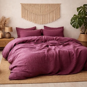 May include: A bedroom with a bed dressed in a deep magenta duvet and pillows. A woven rug and macrame wall hanging add texture. Potted plants and a woven ottoman complete the scene. The bed is approximately 150cm wide.