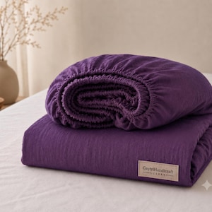 May include: A deep purple fitted sheet with a textured fabric surface, folded neatly. A rectangular beige fabric label is stitched to the bottom corner, featuring the black printed text GaytriHandicraft LUXURY with a horizontal line above and below.