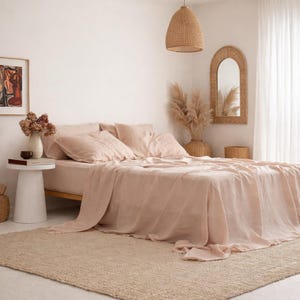 Könnte beinhalten: Ein Schlafzimmer mit einem Bett, das mit rosafarbenen Leinenbettwäsche bezogen ist, einschließlich Spannbettlaken, Bettlaken und Kissenbezügen. Der Raum hat eine neutrale Farbpalette mit einem gewebten Teppich, einem weißen Beistelltisch und einer gewebten Pendelleuchte.