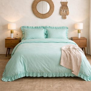 May include: A mint green bedspread with ruffled edges and matching pillowcases. The bed is flanked by two wooden nightstands with lamps. A beige throw blanket rests on the bed, and a woven mirror and wall hanging are above the bed.