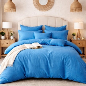 Puede incluir: Un juego de cama azul brillante con almohadas y funda nórdica a juego. Una manta color crema y un libro abierto descansan sobre la cama. Dos mesitas de noche de madera con lámparas flanquean la cama. La habitación tiene una paleta de colores neutros.