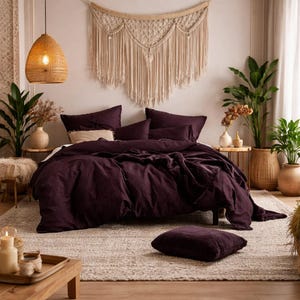 Può includere: Una scena di camera da letto con un letto con biancheria da letto viola scuro, tra cui un copripiumino, cuscini e un cuscino da pavimento abbinato. La stanza è decorata con piante, una lampada a sospensione intrecciata e un arazzo in macramè.