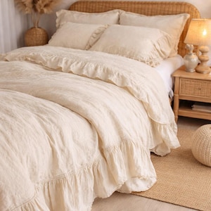 May include: Cream-colored bedding set with ruffled edges, including a duvet cover and pillowcases. The bed has a woven headboard and is next to a wooden nightstand with a lamp. A woven rug and decorative vase are also visible.