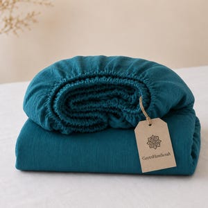 May include: A folded teal fitted sheet with a textured fabric surface. An elasticized edge is visible along the top. A rectangular beige paper tag is attached with a string, featuring a floral mandala icon and the text GaytriHandicraft.