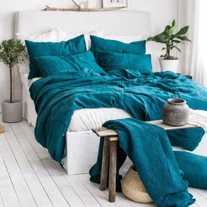Peut inclure: Un ensemble de literie turquoise orne un lit blanc. Un banc en bois supporte une couverture assortie. La pièce présente des murs et un sol blancs, avec des plantes en pot. Un vase aux tons neutres est posé sur le banc.