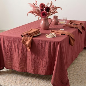 Puede incluir: Un mantel de lino color terracota sobre una mesa, con servilletas de lino plegadas a juego, una jarra de cerámica, un jarrón pequeño, un vaso transparente, conchas marinas y un ramo de flores secas sobre la superficie.