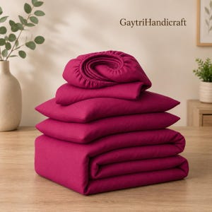 May include: A stack of folded magenta bedding items, including a fitted sheet with visible elastic edges, resting on a wooden surface. The text GaytriHandicraft is printed in the upper right corner of the image above the fabric stack.