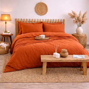 以下が含まれることがあります： オレンジ色の寝具が特徴の寝室のシーン。布団カバーと枕カバーが含まれています。木製のヘッドボードとランプ付きの小さな木製テーブルが見えます。織りのラグと装飾品が、居心地の良いボヘミアンな美学を完成させています。