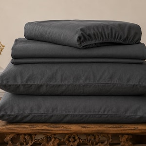 May include: A stack of dark charcoal gray bedding consisting of a fitted sheet, a flat sheet, and two pillowcases. The items are folded neatly in a pile, showcasing a matte, solid-colored fabric texture throughout the entire set of linens.