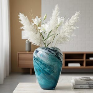 Op de afbeelding: Een decoratieve glazen vaas met een wervelend blauw en wit patroon, gevuld met wit pampasgras en witte lelies. De vaas is een pronkstuk op een tafel met marmeren blad en creëert een moderne en elegante esthetiek.