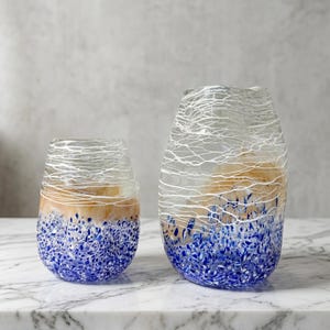 Può includere: Due vasi decorativi in vetro. I vasi presentano una base maculata blu, una sezione centrale beige e un motivo bianco a forma di ragnatela nella parte superiore. I vasi sono di diverse dimensioni e sono appoggiati su una superficie di marmo.