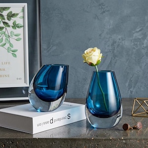 Può includere: Due vasi di vetro blu con base trasparente. Un vaso contiene una rosa gialla. I vasi sono su un libro bianco con la parola "passion" stampata. Un quadro incorniciato e decorazioni geometriche dorate sono sullo sfondo.