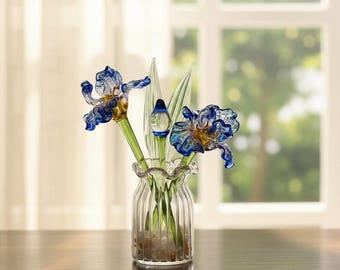 Könnte beinhalten: Eine Glasvase mit Glas-Irisblüten. Die Blüten sind blau und goldfarben, mit grünen Stielen und Blättern. Die Vase steht auf einer Holzoberfläche, im Hintergrund ist ein Fenster zu sehen. Die Vase ist mit kleinen Steinen gefüllt.