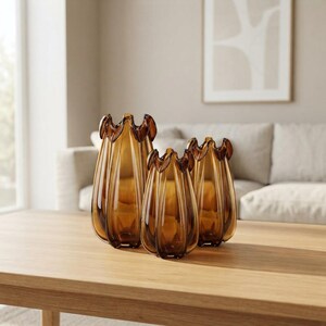 Può includere: Un set di tre vasi in vetro color ambra con finitura traslucida. Ogni vaso presenta una distinta texture a coste verticali e un bordo smerlato e ondulato. I vasi variano in altezza e sono disposti in un gruppo su una superficie piana.