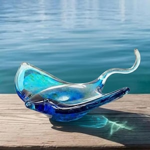 Könnte beinhalten: Eine handgefertigte Glasschale in Form eines Mantarochens. Die Schale besteht aus klarem, blauem und türkisfarbenem Glas mit einem gebogenen Schwanz und flügelartigen Seiten. Die Schale steht auf einer Holzoberfläche mit verschwommenem Wasserhintergrund.