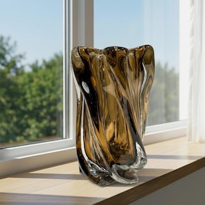 May include: A decorative brown glass vase with a unique, twisted design. The vase has a clear base and a dark brown, swirling pattern that creates a textured appearance. It sits on a wooden windowsill, with a window and trees in the background.