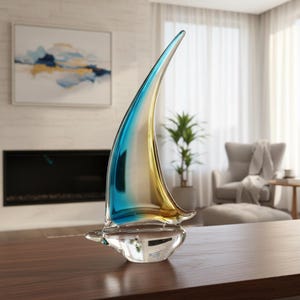 Può includere: Una scultura di barca a vela in vetro trasparente con una vela blu e oro. La scultura poggia su una base trasparente ed è esposta su una superficie di legno marrone scuro. La vela è curva e presenta una sfumatura di colori.