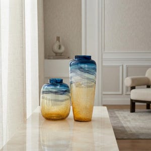 Può includere: Due vasi in vetro con un design sfumato che passa da una parte superiore blu intenso a una base ambrata testurizzata. Il vaso più alto è cilindrico, mentre quello più basso è arrotondato. Entrambi presentano linee ondulate orizzontali.