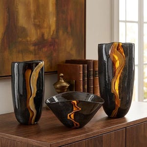 May include: Three decorative glass vases and a bowl in a modern style. The vases are black with a flowing amber and gold design. The bowl is black with a similar amber and gold design. The objects are displayed on a wooden surface.