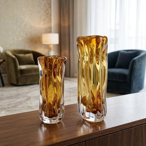 Può includere: Due vasi di vetro color ambra con un design strutturato e ondulato. I vasi sono di dimensioni diverse e poggiano su una superficie di legno scuro. Il vaso più grande è più alto di quello più piccolo. Il vetro ha una finitura lucida.