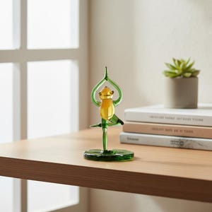 Può includere: Una statuetta di rana di vetro in posa yoga. La rana è gialla e verde, con le braccia alzate sopra la testa. Si trova su una base circolare verde. Una piccola pianta in vaso e libri sono sullo sfondo.