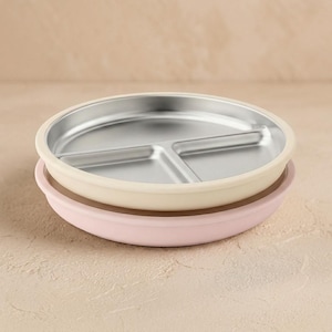 May include: Two stacked round divided plates with a metallic silver interior and smooth silicone rims. The top plate features a cream-colored rim, while the bottom plate has a soft pastel pink rim. Each plate is divided into three sections.