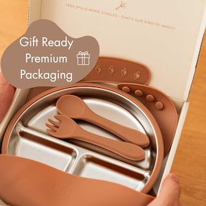 May include: A baby feeding set in a gift-ready box. The set includes a stainless steel divided plate, a spoon, a fork, and a bib, all in a warm terracotta color. The box has the text "LESS SPILLS MORE GIGGLES - THAT'S OUR KIND OF MAGIC!" and "Gift Ready Premium Packaging."