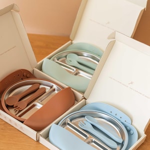 May include: Three boxed baby feeding sets. Each set includes a stainless steel divided plate, a silicone bib, and a fork and spoon. The bibs and utensils are in shades of brown, blue, and green. The boxes have text that reads "Less spills more giggles - That's our kind of magic!"