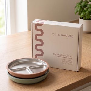 May include: A set of two stainless steel plates with suction bases. The plates are divided into three sections and sit on a green and a brown silicone base. The box reads "TOTS GROUND TINY BITES DUO PLATE SET".