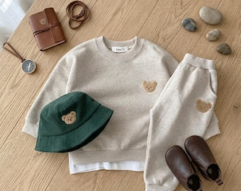 May include: A beige long-sleeve sweatshirt with a small embroidered bear, matching beige sweatpants with a bear patch, a dark green corduroy bucket hat with a bear patch, and brown leather-like shoes. The sweatshirt tag reads Size 73.