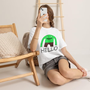 Puede incluir: Una camiseta blanca de manga corta con un gráfico de una criatura verde de brazos largos que lleva pantalones cortos marrones, situado sobre la palabra HELLO en letras mayúsculas negras. La camiseta se lleva con pantalones cortos de mezclilla gris.