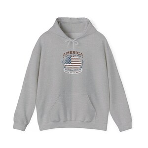 Op de afbeelding: Een lichtgrijze hoodie met een voorzak en een capuchon met trekkoord. De voorkant heeft een cirkelvormige afbeelding met de Amerikaanse vlag, het woord "AMERICA" en de zinnen "LAND OF THE FREE" en "HOME OF THE BRAVE".