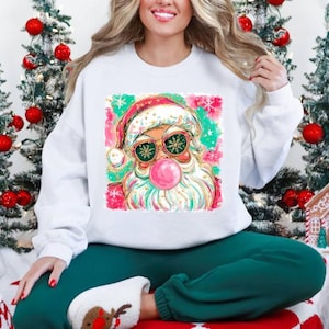 May include: White sweatshirt featuring a colorful graphic of Santa Claus wearing sunglasses and blowing a bubble. The Santa graphic has pink, green, and gold accents. The person is wearing green sweatpants and reindeer slippers. Christmas trees and decorations are in the background.