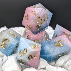 Könnte beinhalten: Ein Set aus sieben Würfeln mit einem Farbverlauf von Rosa und Blau. Die Würfel haben goldfarbene Zahlen und liegen auf weißen Steinen. Diese Würfel sind für Tischrollenspiele geeignet.