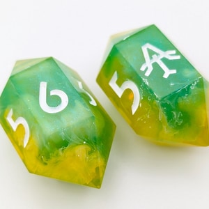 Könnte beinhalten: Zwei polyedrische Würfel mit grünem und gelbem Marmoreffekt. Die Würfel haben weiße Zahlen und Symbole. Die Würfel werden für Tischspiele und Rollenspiele verwendet.