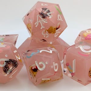 Könnte beinhalten: Ein Satz von sieben polyedrischen Würfeln aus rosa, durchscheinendem Harz. Jeder Würfel hat weiße Zahlen und farbenfrohe Illustrationen zum Thema Essen, darunter Donuts und Cupcakes. Die Würfel sind für Tischspiele konzipiert.