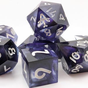 Könnte beinhalten: Satz von Würfeln mit einem dunkelblauen und violetten Marmoreffekt. Die Würfel haben silberne Zahlen und sind auf weißem Hintergrund angeordnet. Das Set enthält verschiedene Formen, darunter ein D20, D12, D10, D8 und D6.